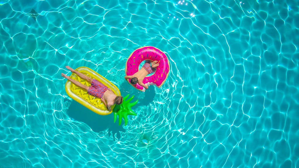 Two kids enjoying a sunny day on colorful floaties in a turquoise swimming pool.